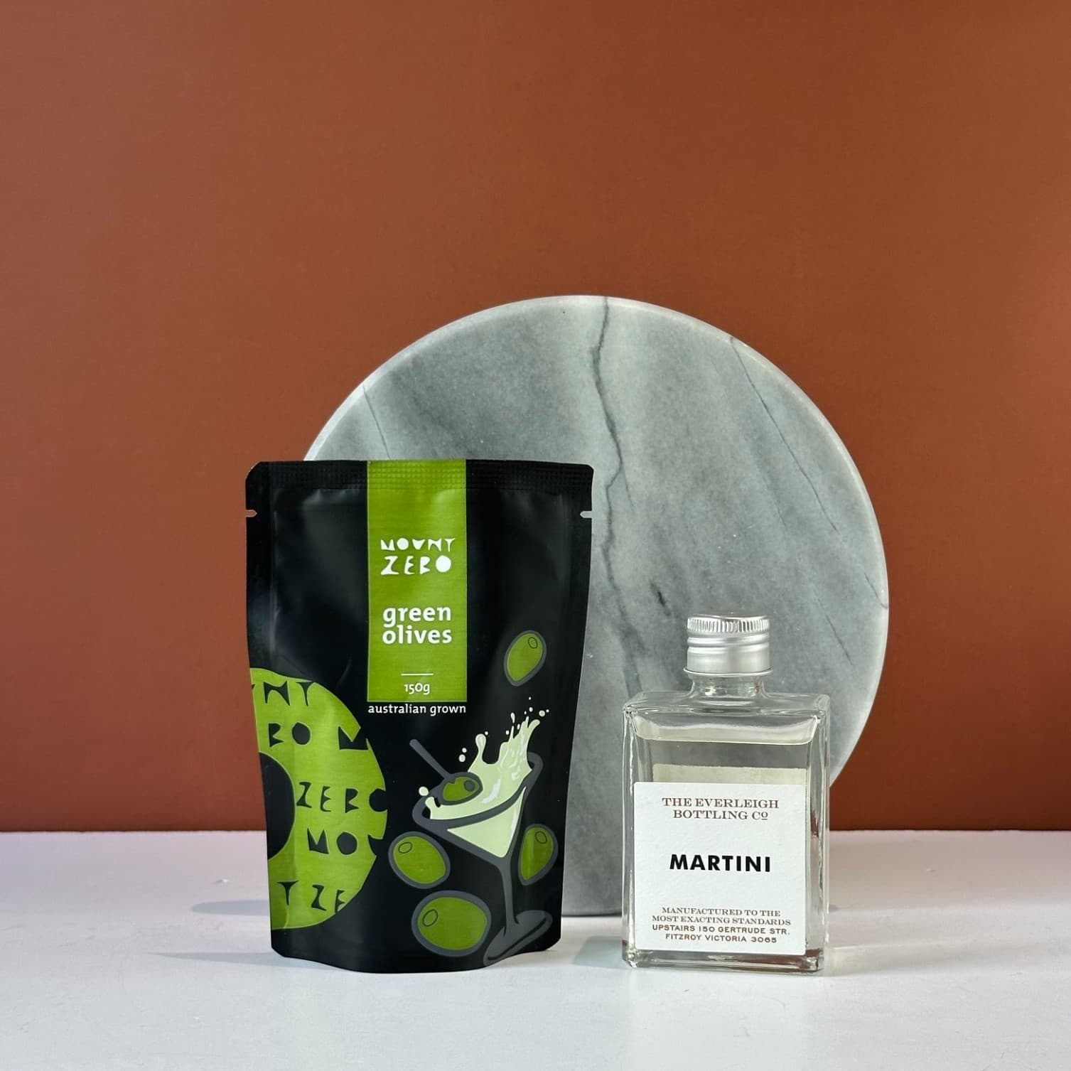 The Mini Happy Hour Classic Martini gift set elegantly displayed featuring The Everleigh Bottling Co. Martini in a minimalist glass bottle and Mount Zero green olives in contemporary black and green packaging, arranged on a marble surface against a sophisticated terracotta backdrop, representing a premium cocktail experience